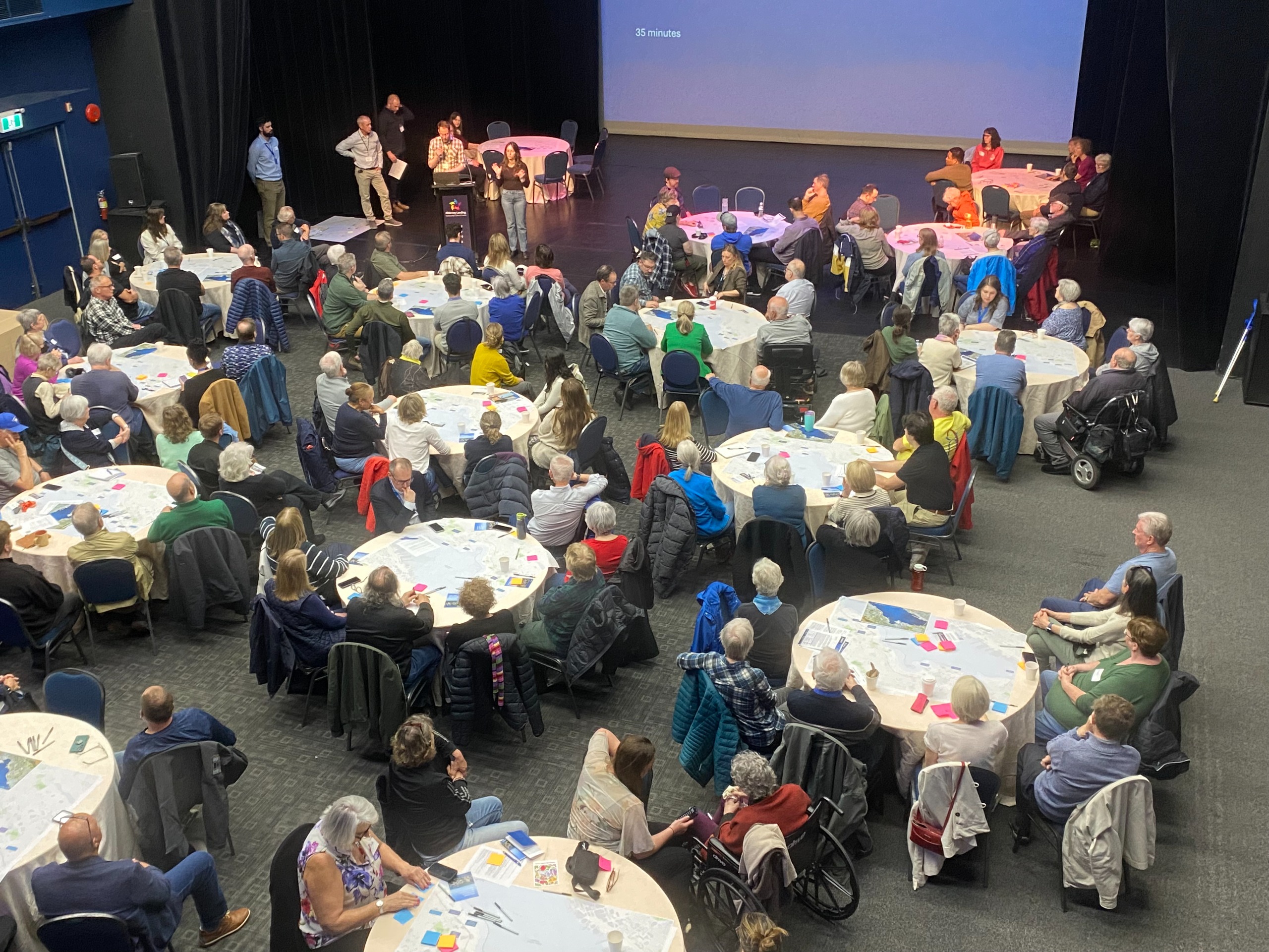 An engagement session at Alderney Landing Theatre. There are presentation on the stage with multiple round tables with people doing activities and listening to presenters.